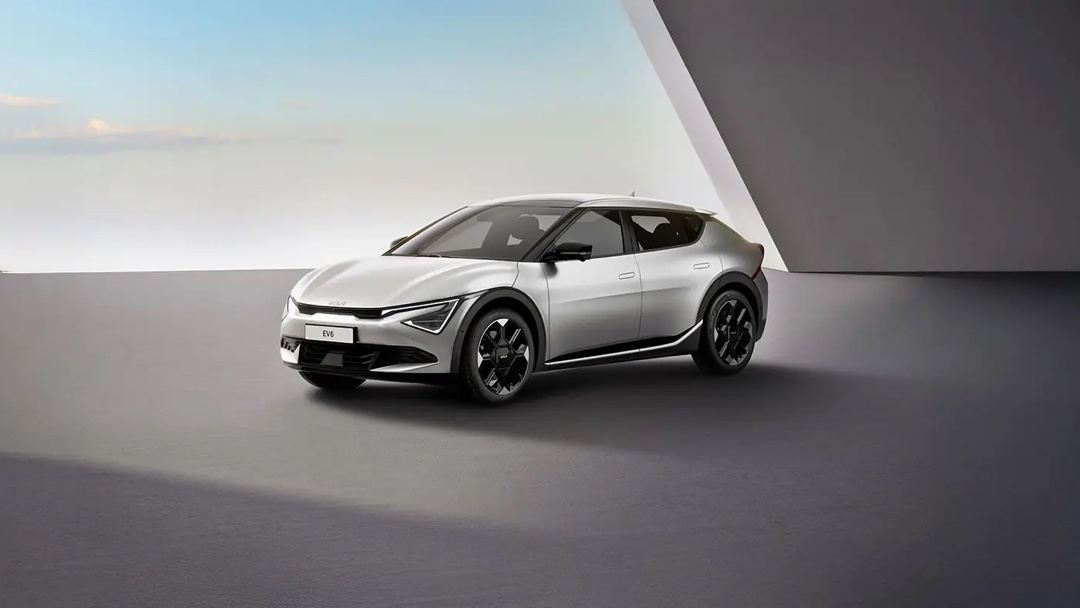 Kia EV6 2025 en gris métallisé, vue trois quarts avant, stationnée sur une surface minimaliste avec fond architectural.